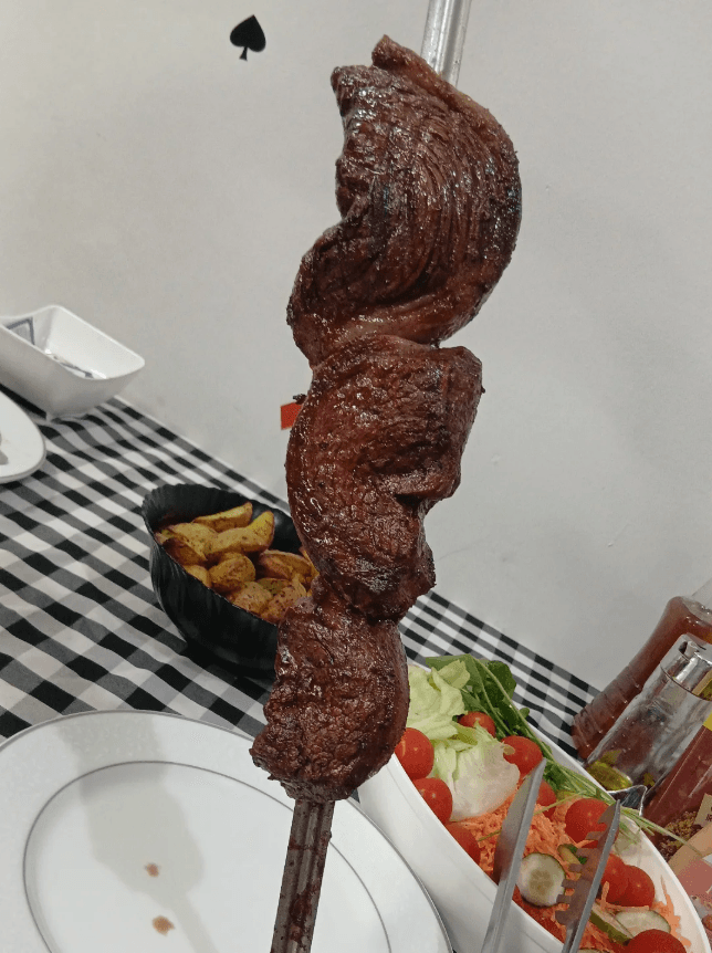 Espeto de churrasco com dois cortes de carne, ao lado de batata assada e saladas