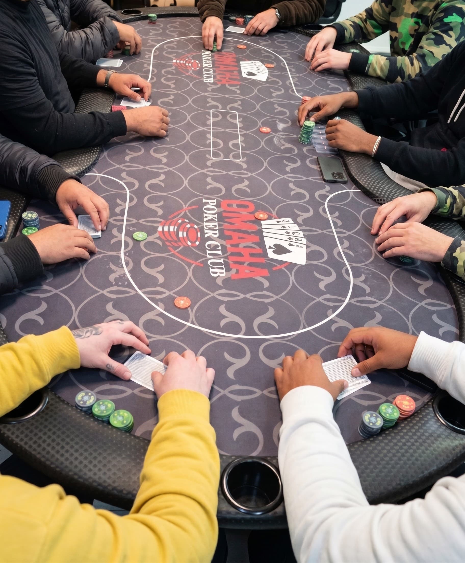 Mesa cheia numa noite do clube, vista de cima: dez pares de mãos, fichas, cartas e a logo estampada no feltro