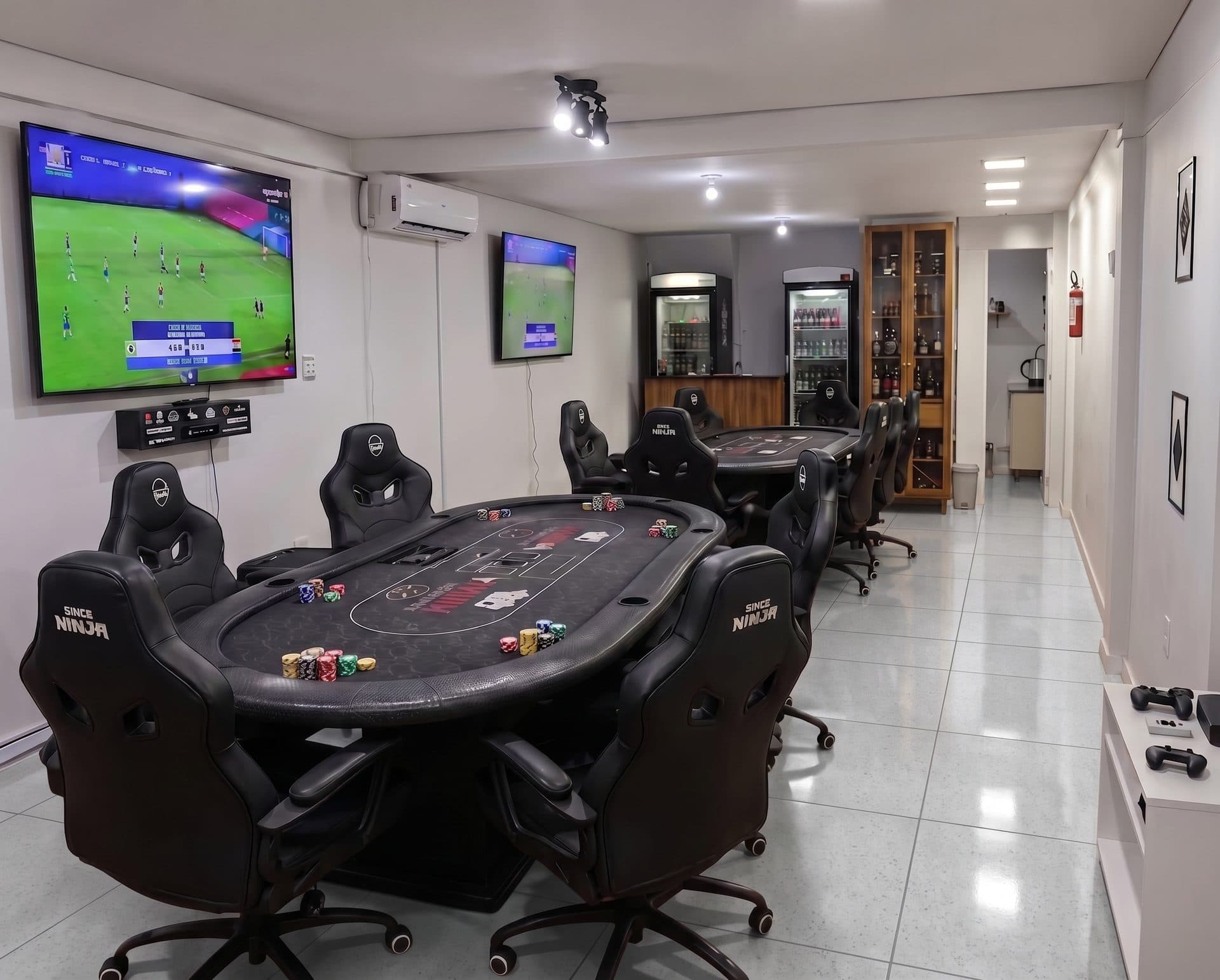 O salão do clube: duas mesas de poker montadas com fichas, poltronas gamer, duas TVs grandes e o bar ao fundo