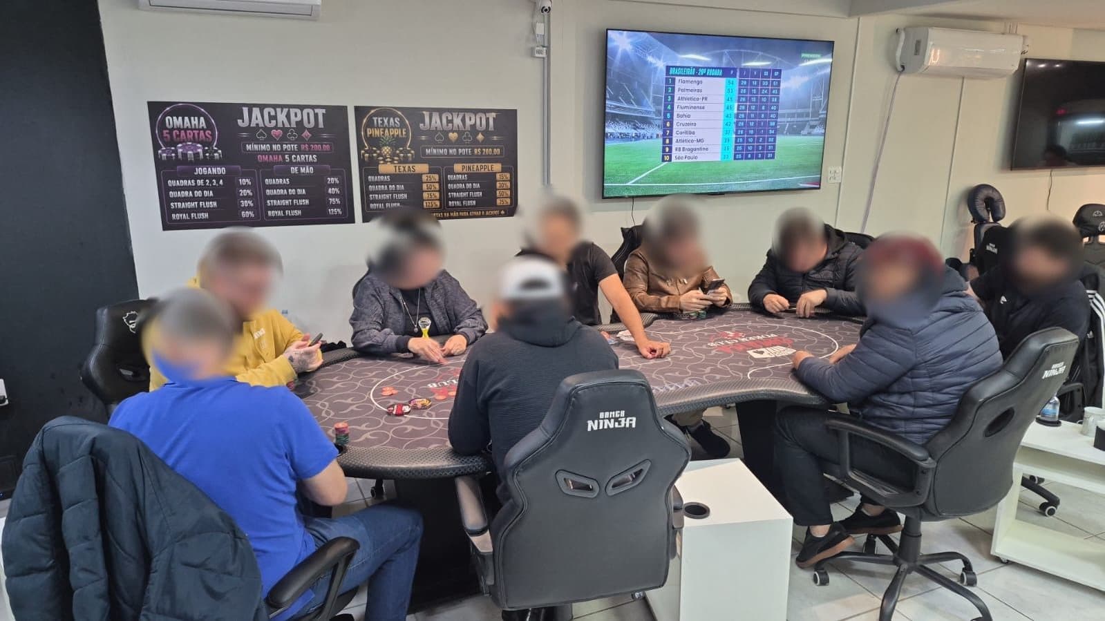 Mesa de Omaha cheia numa noite do clube, com os dois quadros de jackpot na parede e a TV ligada ao fundo